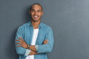 Portrait of handsome man standing with arms crossed in smart casual clothing against a grey wall. Smiling young african guy looking at camera leaning on grey background with copy space. Portrait of a satisfied man looking at camera.