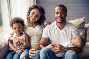 Portrait of beautiful young Afro American parents and their cute children looking at camera and smiling while sitting on sofa at home. Little baby is sleeping in dad's arms